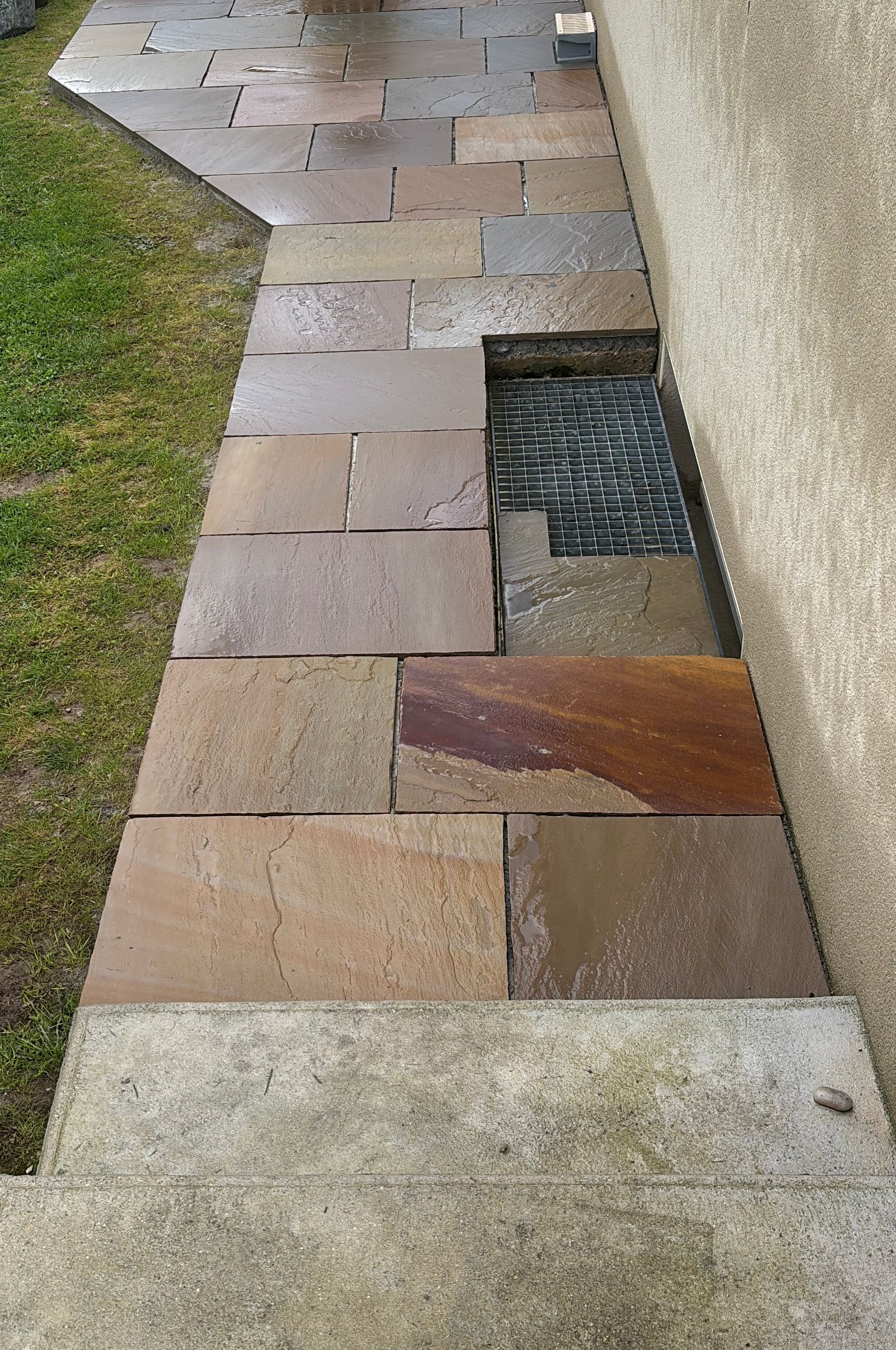 Allée en pierre avec des pavés inégaux et une section manquante laissant apparaître un drain à côté d'un mur et d'une pelouse.