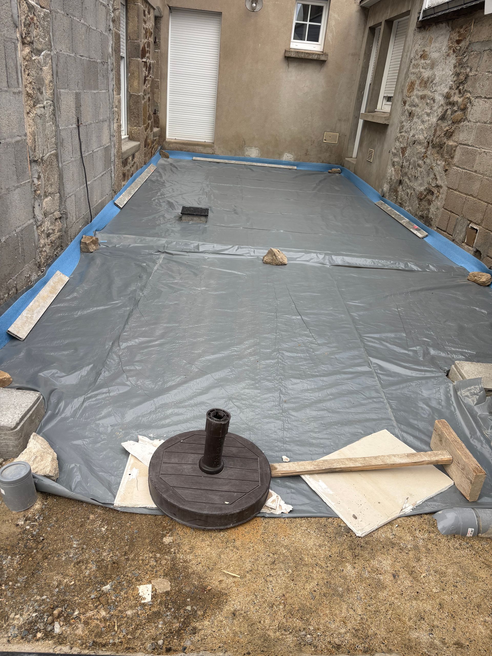 Cour intérieure couverte d'une bâche en plastique, avec un tuyau de drainage et des murs en pierre tout autour.