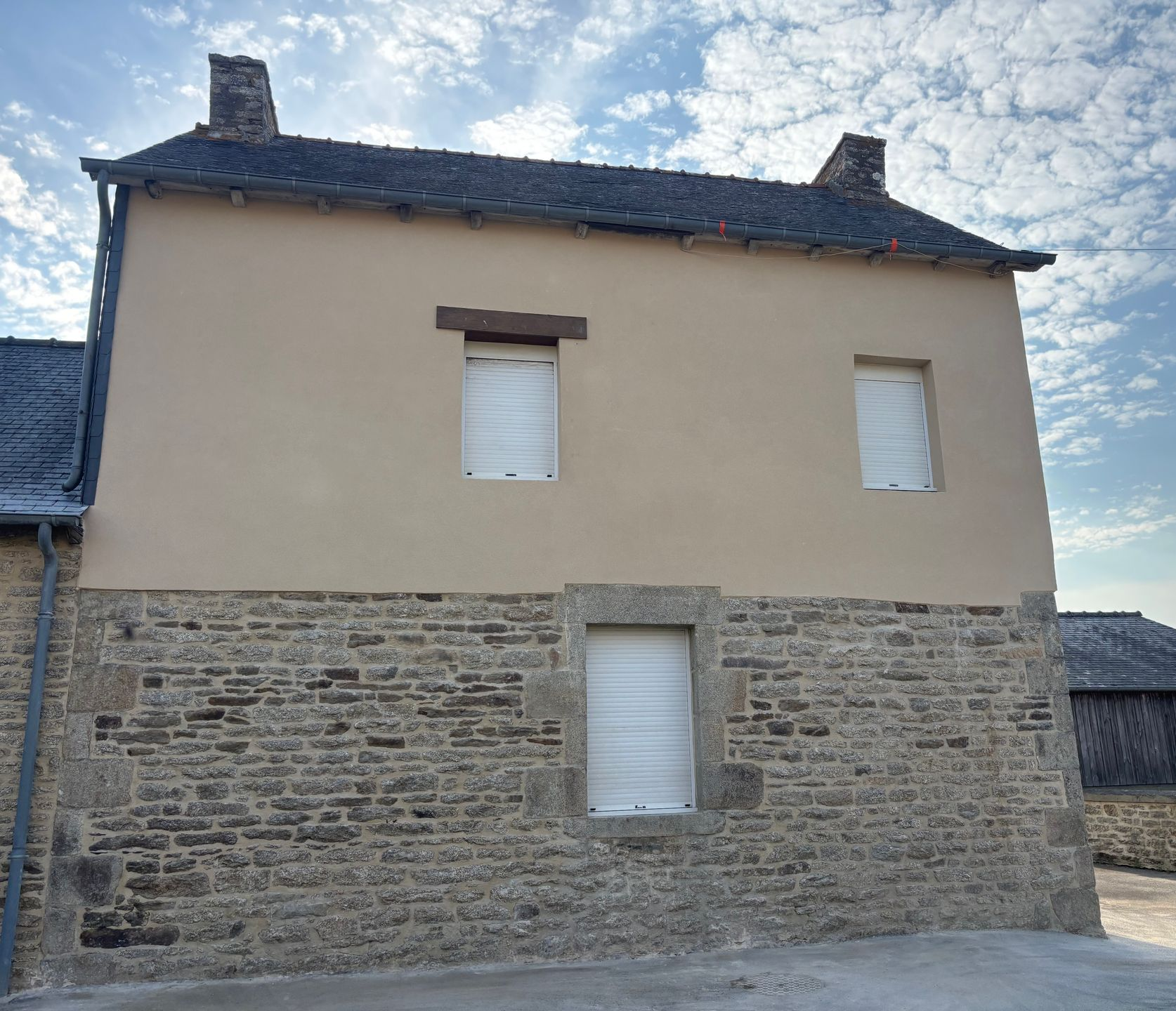 Maison en pierre et plâtre avec un toit en ardoise et des fenêtres à volets sous un ciel nuageux