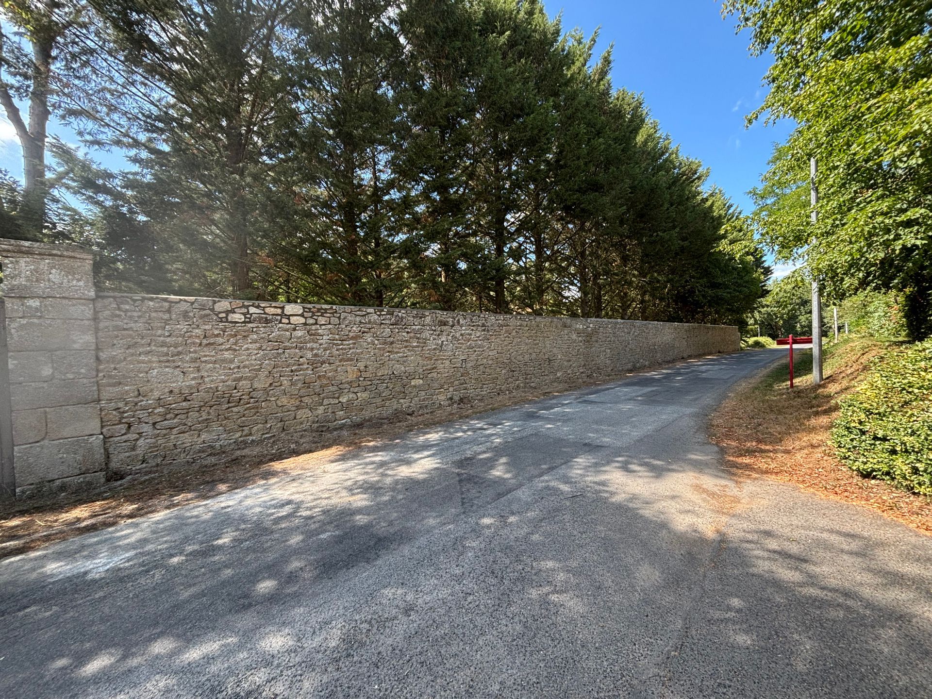 Un mur de pierre longe une allée de gravier sinueuse bordée d'arbres, sous un ciel bleu limpide.