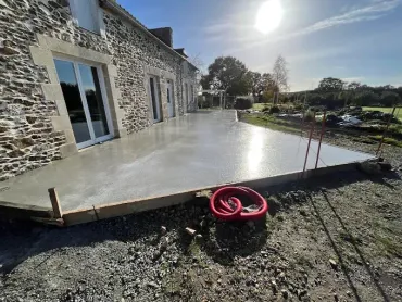 Nouvelle terrasse en béton à côté de la maison en pierre, avec un enrouleur de tuyau d'arrosage au premier plan et un jardin ensoleillé au-delà.