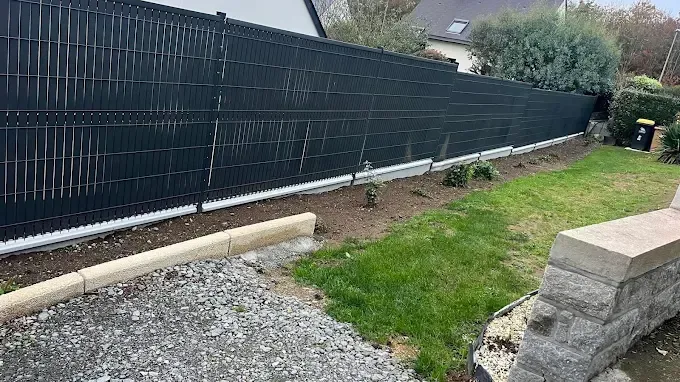 Cour arrière avec clôture noire pour plus d'intimité, chemin de gravier, pelouse et muret de pierre sous un ciel nuageux