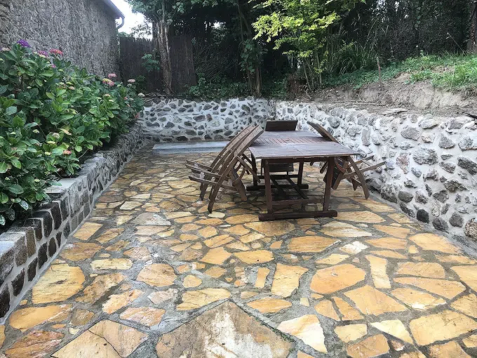 Terrasse en pierre avec une table et des chaises en bois dans une cour de jardin, bordée de murets en pierre et de verdure.