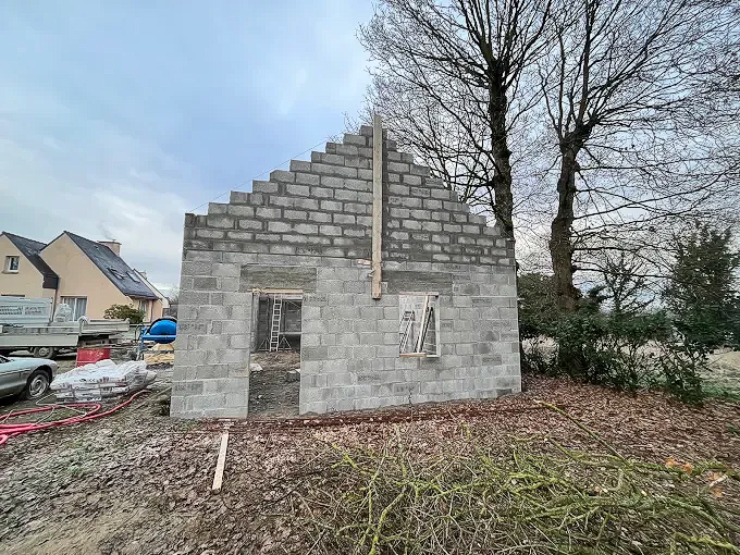 Maison en parpaings partiellement construite dans une cour avec des arbres dénudés et un ciel nuageux