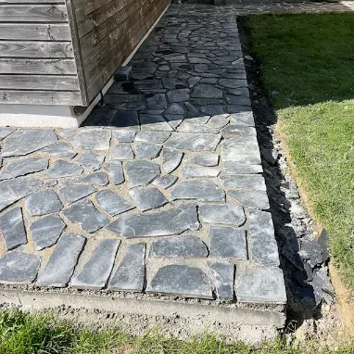 Allée en pierre longeant un bâtiment en bois, bordée de gazon et de dalles de béton.