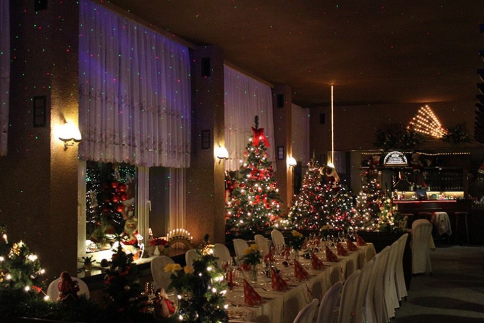 Weihnachtlich dekorierter Bankettsaal mit mehreren beleuchteten Weihnachtsbäumen, einem langen, für ein Essen gedeckten Tisch und Fensterdekorationen.