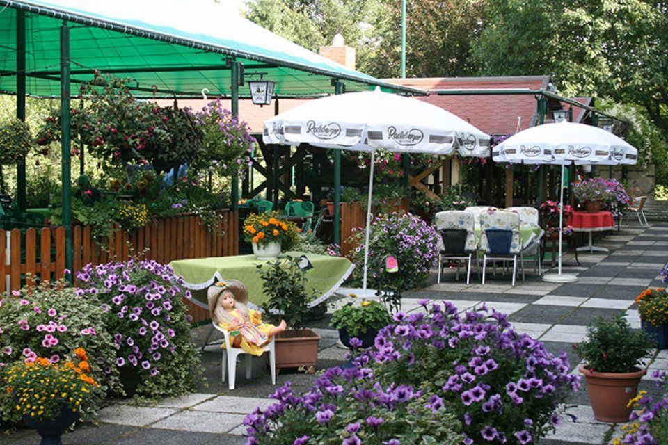 Straßencafé mit Tischen, Sonnenschirmen und hängenden Blumenkörben; eine Puppe sitzt an einem Tisch.
