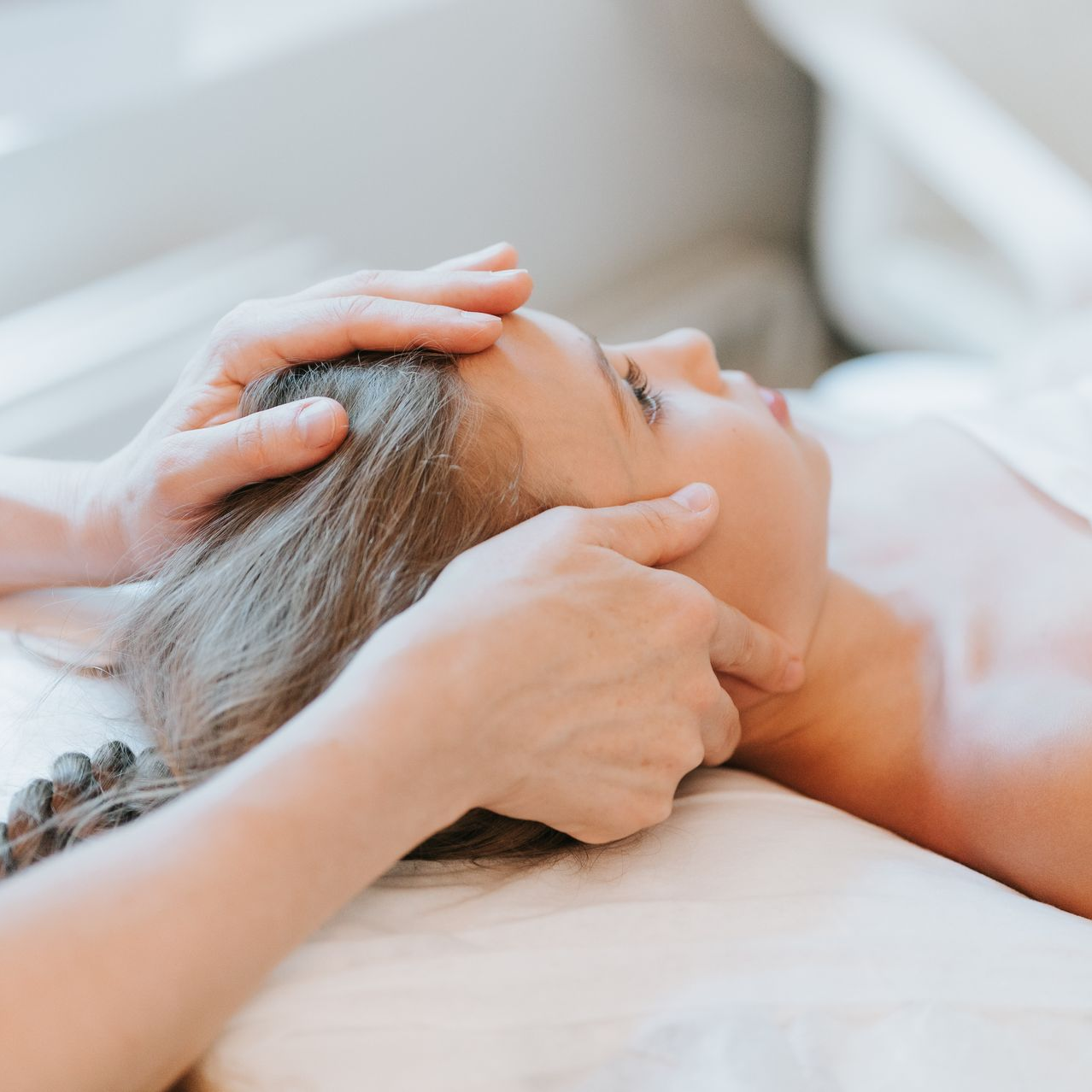 Personne effectuant un massage du visage, les mains sur le front et les tempes d'une femme allongée sur un lit à l'intérieur.