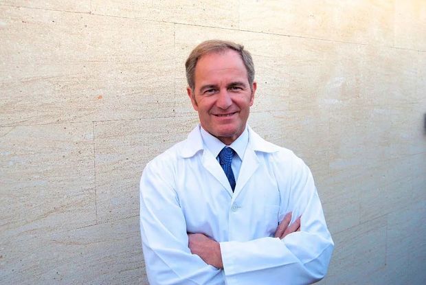 Un hombre con bata de laboratorio y corbata está de pie con los brazos cruzados.