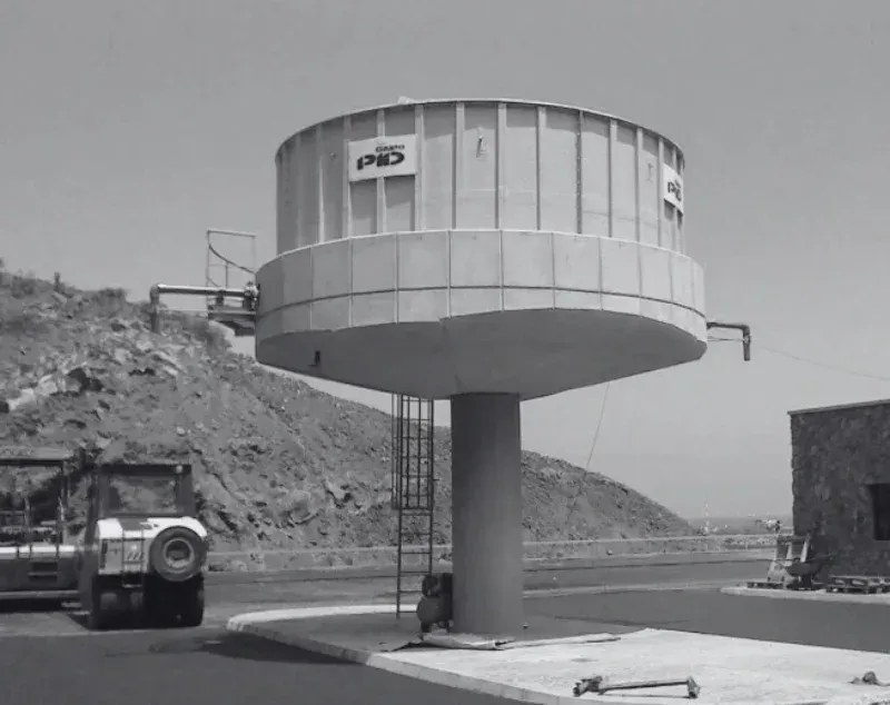 Estructura industrial cilíndrica gris sobre un único pilar junto a una carretera, con un camión y una ladera rocosa cerca.