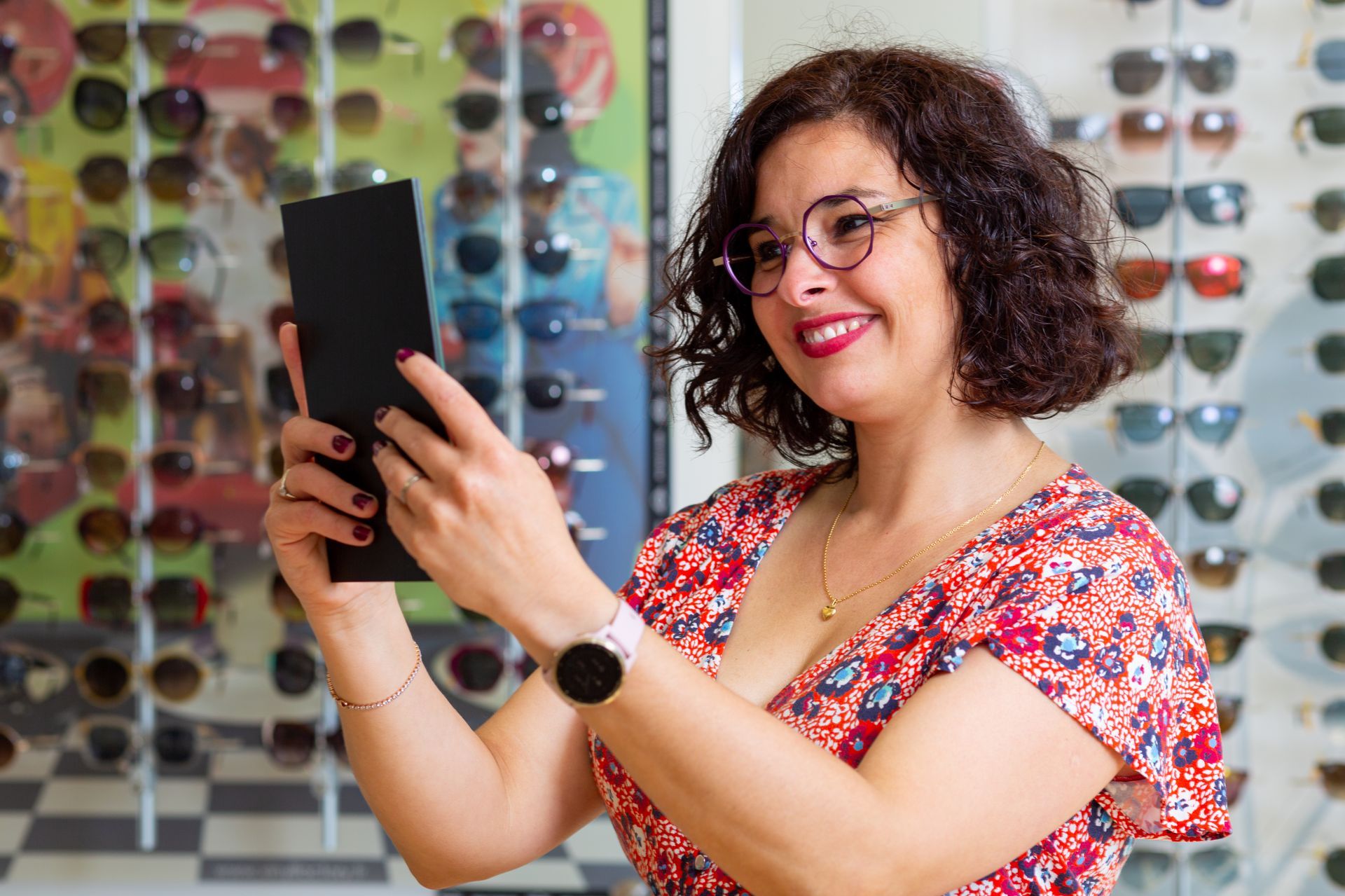 Une femme sourit en tenant un miroir devant un mur de lunettes.