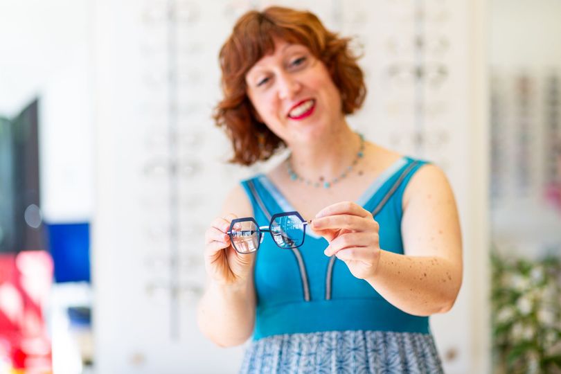 Femme tenant des lunettes bleues dans un magasin lumineux, souriant à l'objectif.