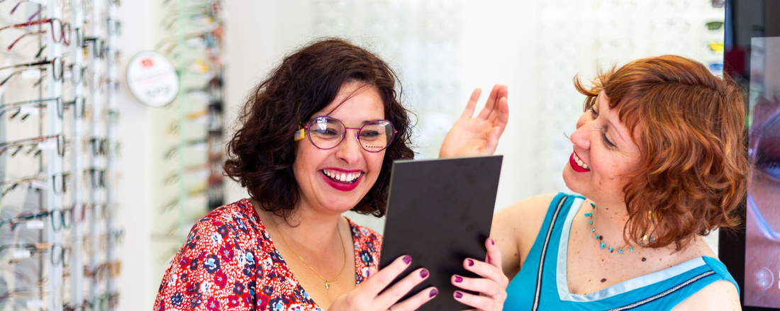 Deux femmes sourient devant une tablette dans une boutique d'optique lumineuse.