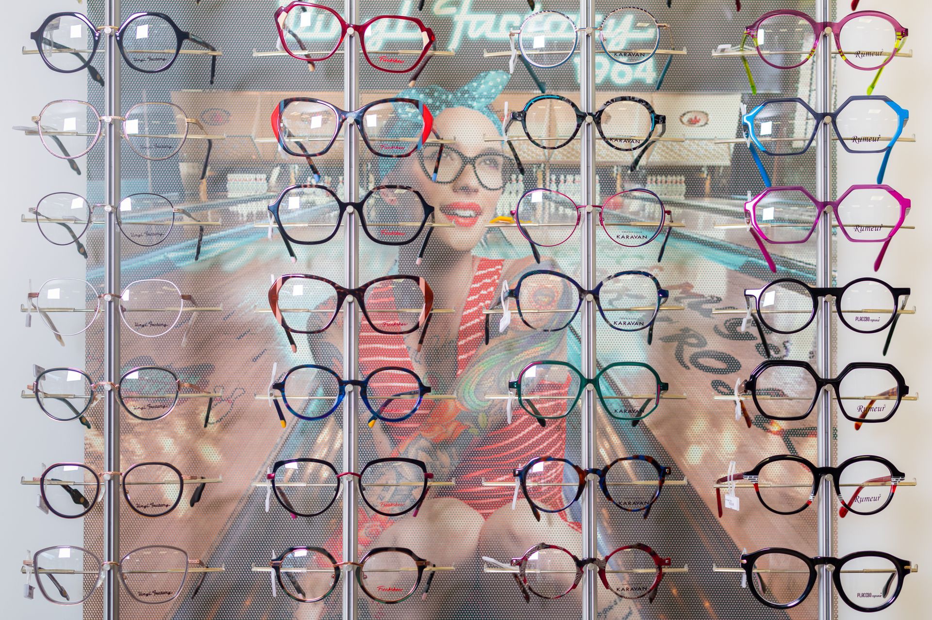 Présentoir de montures de lunettes colorées sur le mur d'un magasin, avec une personne reflétée derrière.