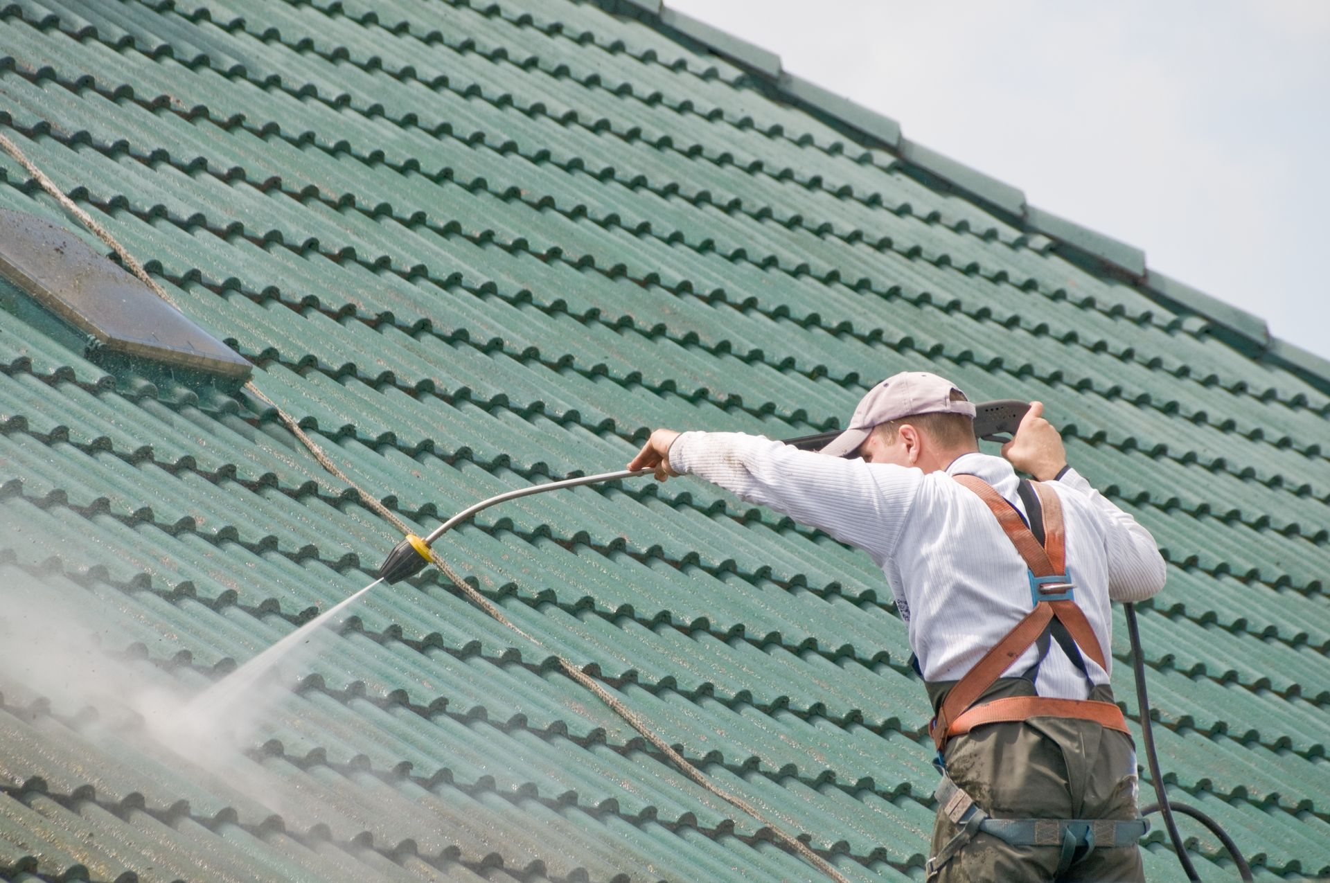 Un homme nettoie à haute pression un toit de tuiles vertes, portant un harnais de sécurité.