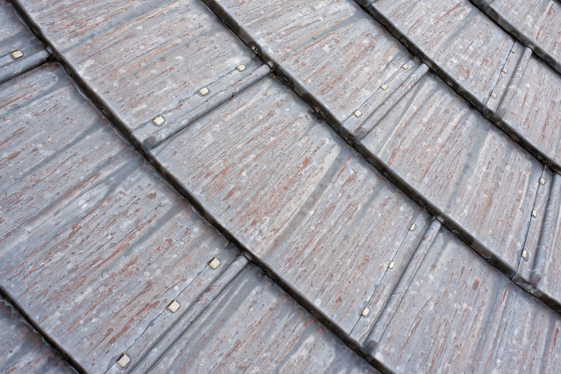 Gros plan sur des tuiles de toit grises patinées, disposées en rangées superposées, montrant leur texture et leurs fixations.