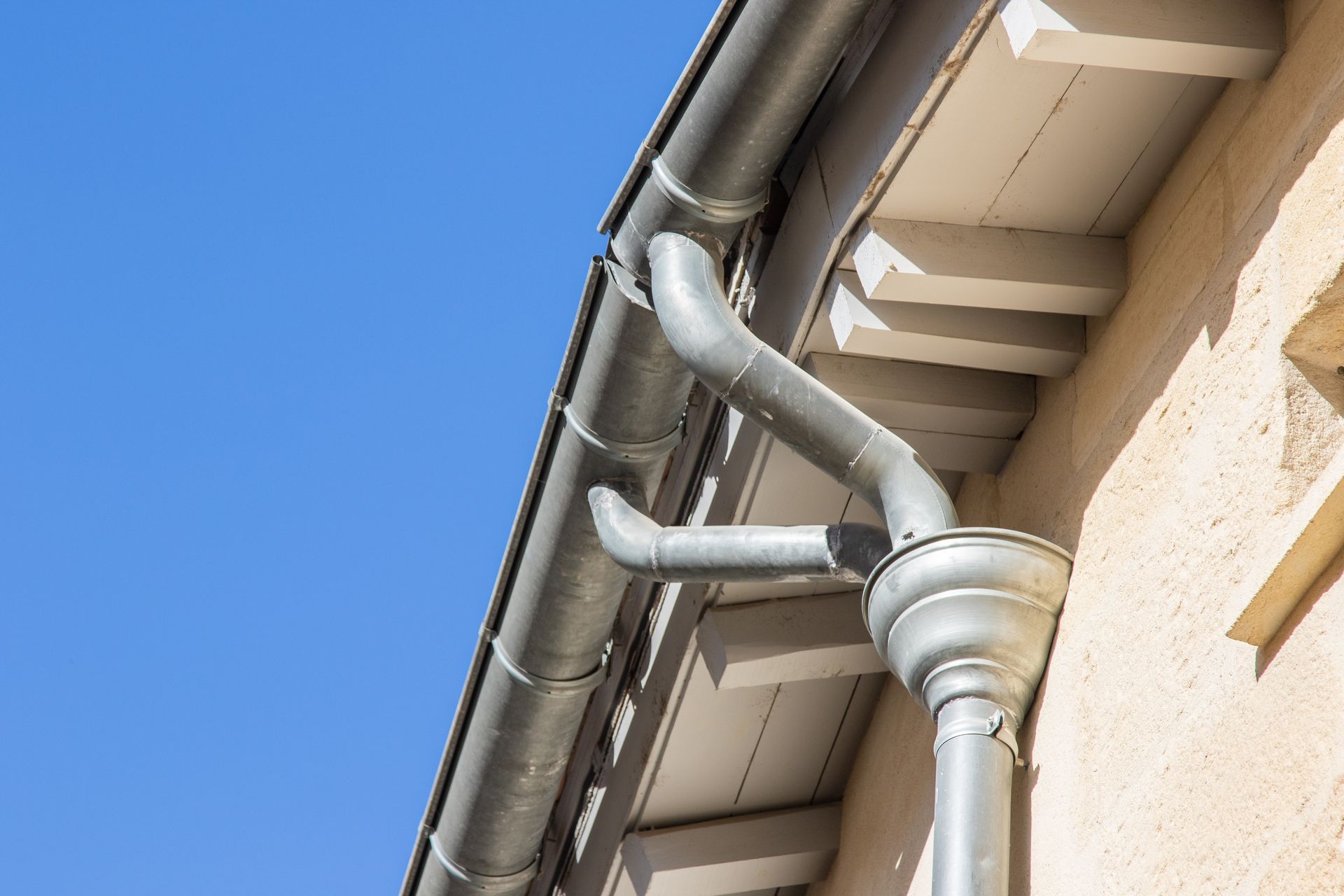 Système de gouttière en métal gris fixé au bord d'un bâtiment beige.