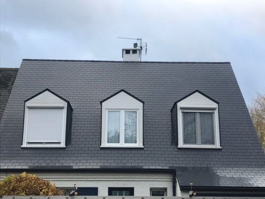 Toit gris avec trois lucarnes, une cheminée et une antenne se détachant sur un ciel nuageux.