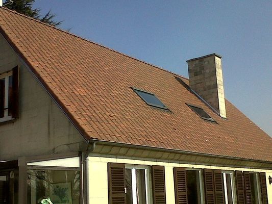 Maison beige au toit de tuiles rouges, avec deux puits de lumière, une cheminée et des volets marrons, se détachant sur un ciel bleu.