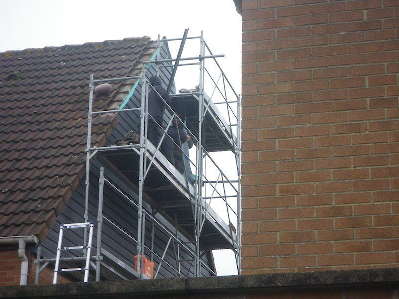 Échafaudages à côté d'un bâtiment en briques et d'un toit ; des ouvriers travaillent sur les échafaudages.