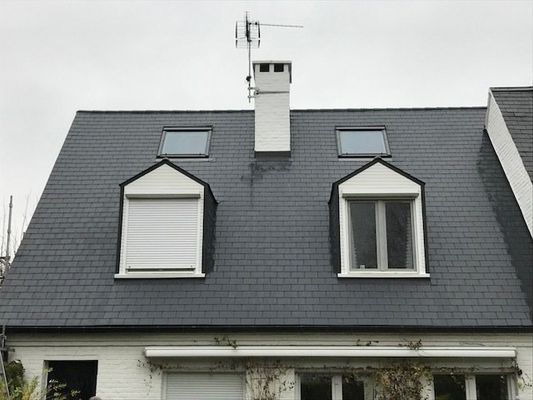 Maison blanche au toit de bardeaux sombres, avec deux lucarnes, une cheminée et une antenne de télévision. 