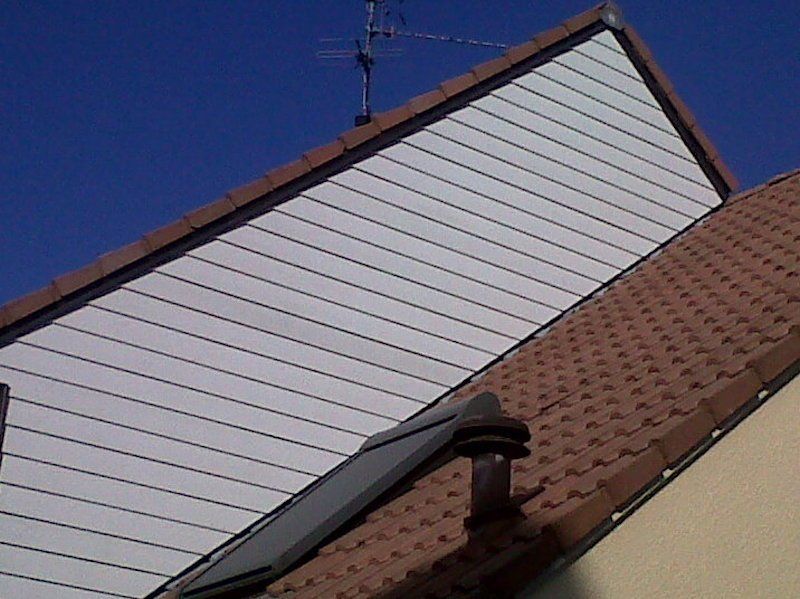 Toit de tuiles brunes et bardage blanc avec une cheminée et une antenne visibles.