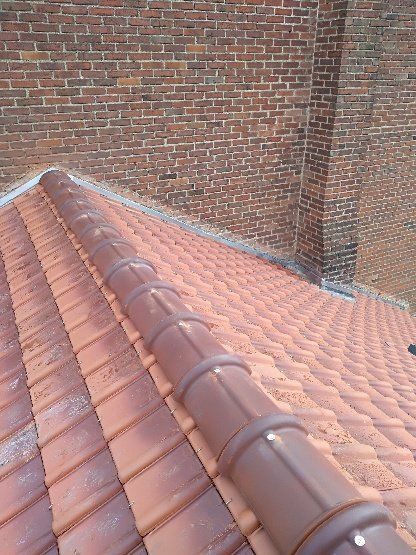 Toit de tuiles rouges avec une faîtière brune, contre un mur de briques brunes.