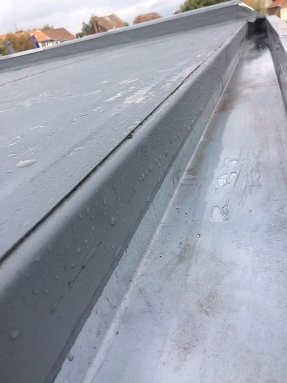 Gros plan sur un toit en métal gris avec un canal, probablement pour l'évacuation de l'eau, laissant apparaître quelques gouttelettes.