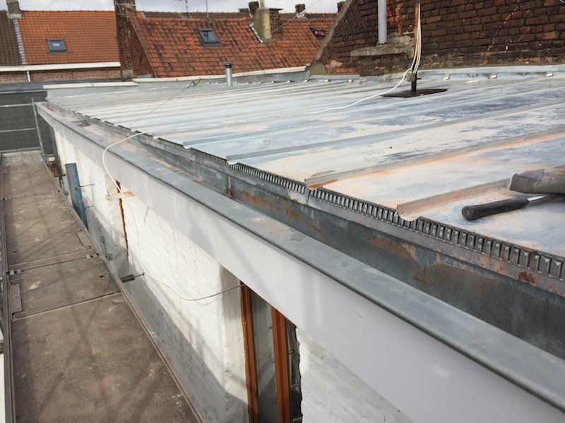 Un toit plat en métal avec une gouttière et un bord de bâtiment ; à l'arrière-plan, d'autres toits.