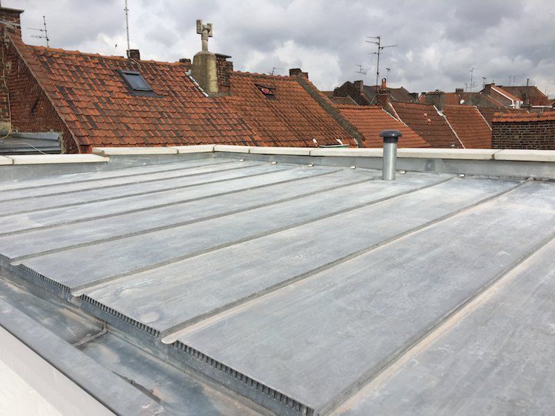 Un toit plat en métal gris aux joints saillants, avec des toits et des bâtiments en tuiles rouges en arrière-plan.