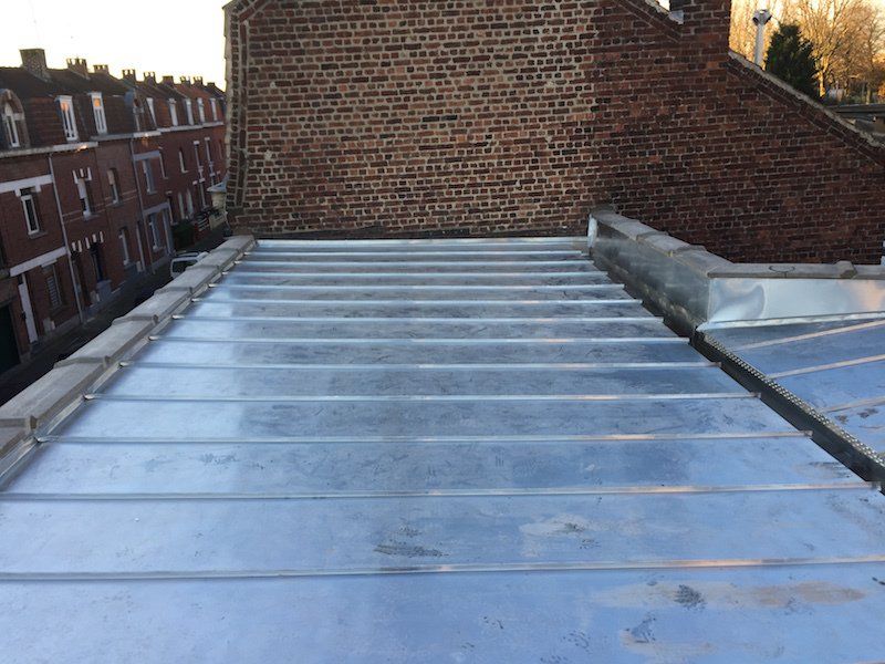 Toiture métallique à panneaux horizontaux contre un bâtiment en briques, autres bâtiments en arrière-plan.