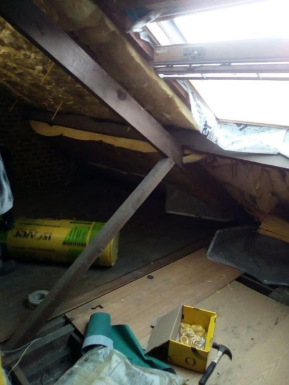 Intérieur du grenier avec poutres apparentes, isolation et puits de lumière. Rouleau d'isolant jaune et divers matériaux visibles.