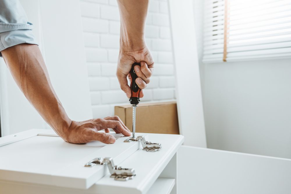 Manos utilizando un destornillador para ensamblar muebles blancos sobre una mesa con herrajes metálicos.