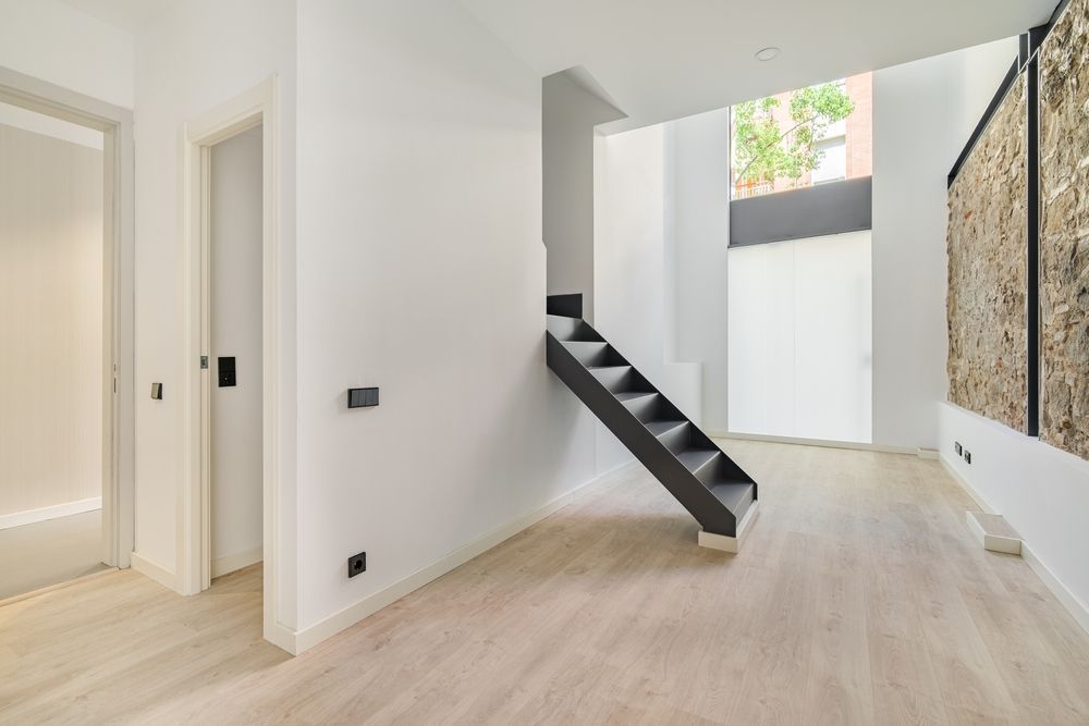 Interior moderno y minimalista con paredes blancas, suelos de madera clara y una escalera negra bajo una claraboya.