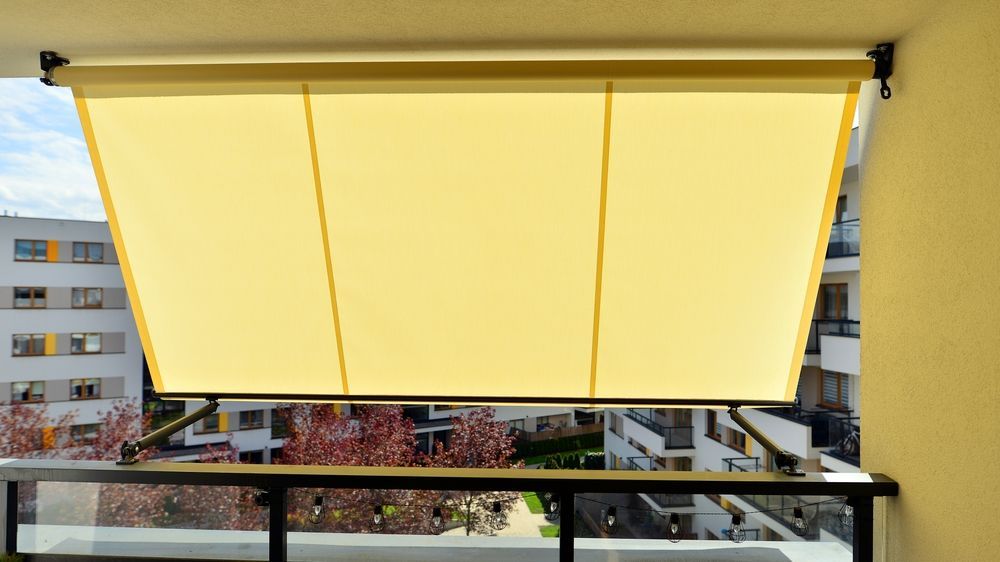 Un toldo amarillo bajado en el balcón ofrece vistas a edificios de apartamentos y árboles.