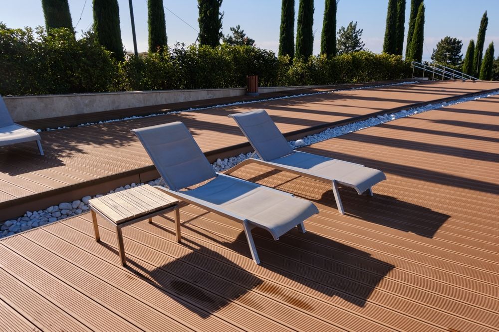 Dos tumbonas grises en una terraza de madera soleada junto a una piscina, con altos cipreses al fondo.