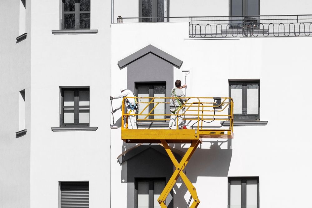 Trabajadores en una plataforma elevadora amarilla pintan la fachada de un edificio blanco.