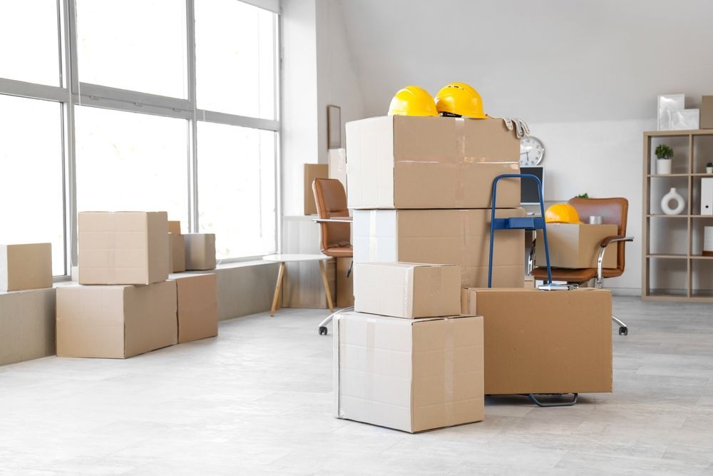 Cajas de mudanza apiladas en una habitación luminosa con dos cascos amarillos encima y una escalera cerca.