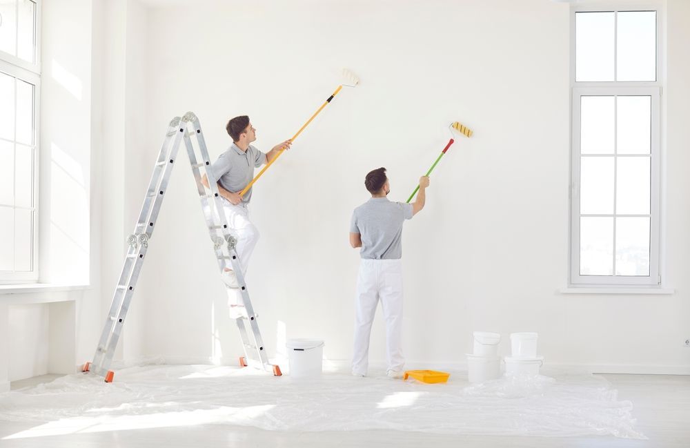 Dos personas pintando una habitación de color blanco brillante, una subida a una escalera y la otra usando un rodillo.
