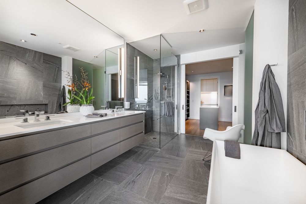 Baño moderno con lavabo doble, ducha de cristal, azulejos grises y un banco de bañera blanco.