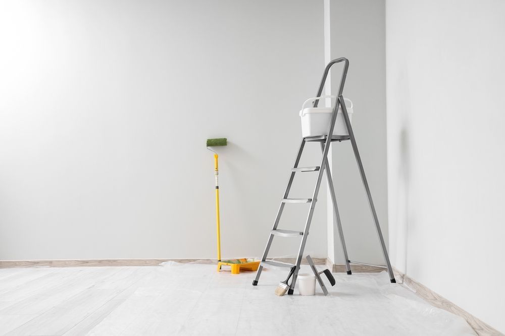 Rodillo de pintura y escalera en una habitación vacía con paredes grises y suelo blanco, sugiriendo trabajo de pintura.