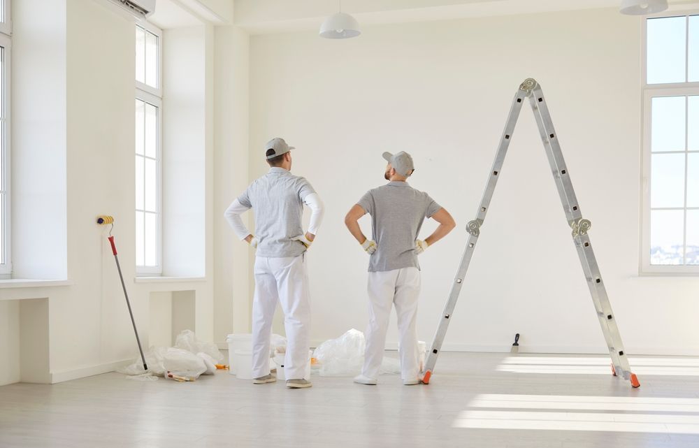 Dos pintores con monos blancos están de pie en una habitación luminosa y vacía, junto a una escalera y herramientas de pintura.