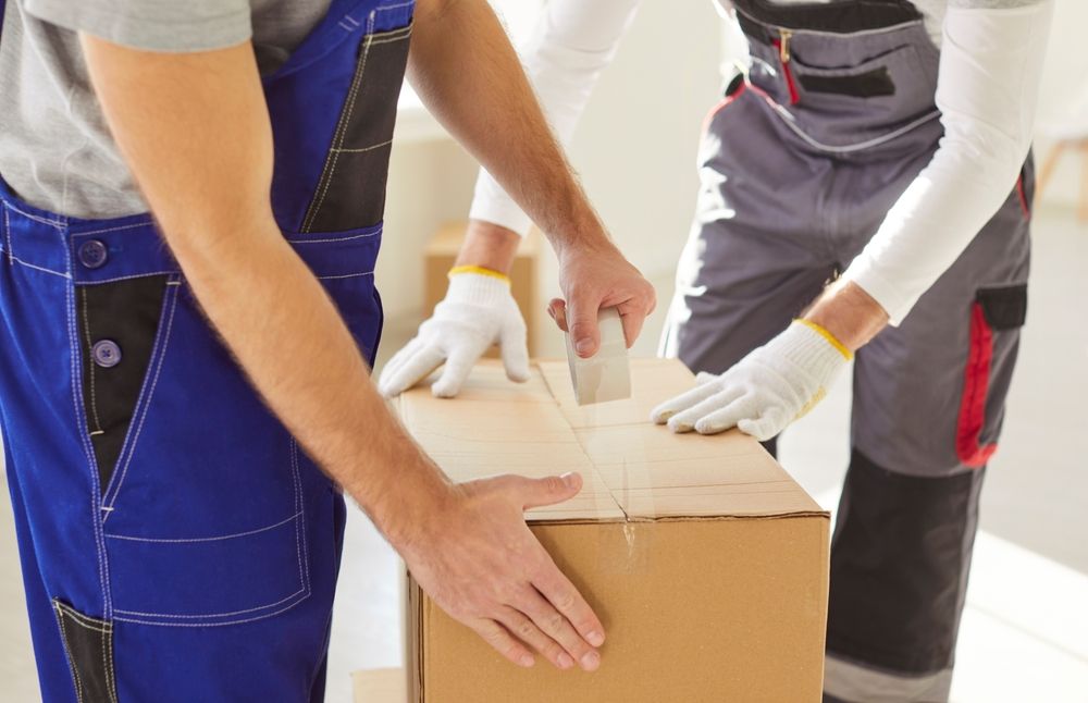 Trabajadores con guantes empaquetan una caja de cartón con cinta adhesiva y documentación.
