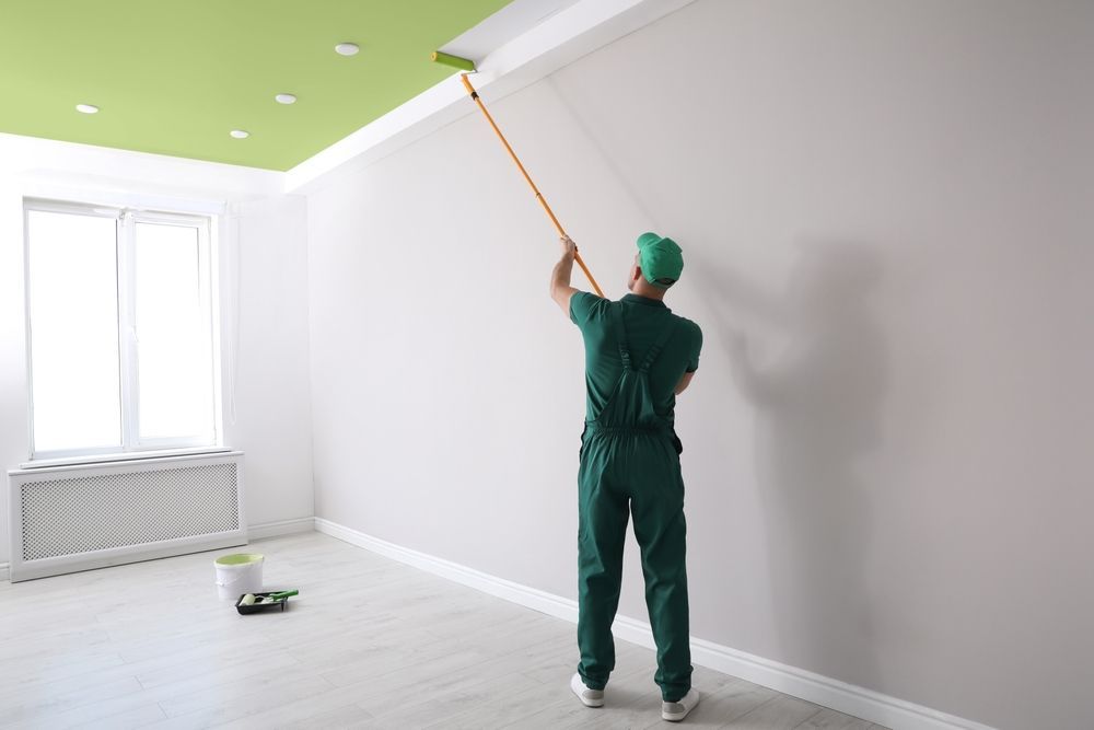 Una persona pinta una pared blanca de una habitación con un rodillo en un palo largo; el techo es verde.