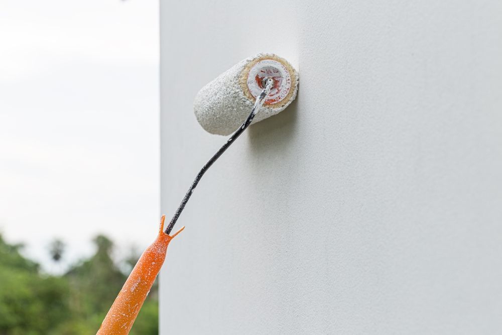 Rodillo de pintura con mango naranja aplicando pintura blanca en la esquina de una pared exterior.