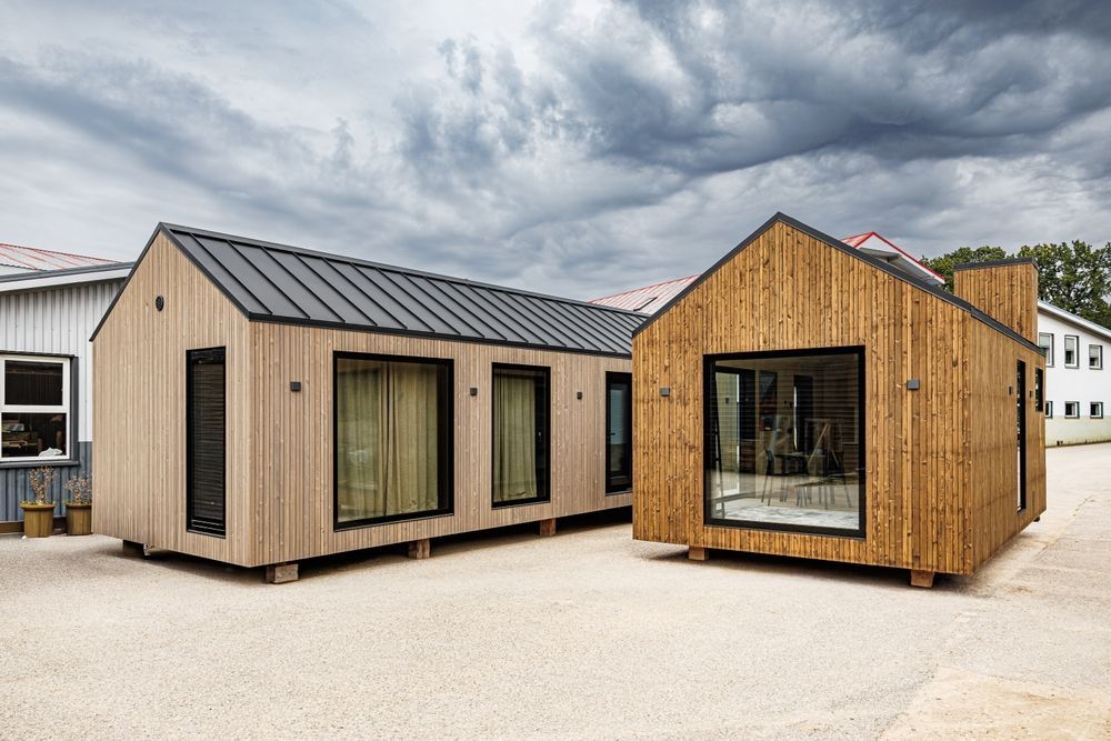 Dos pequeñas casas modernas en un terreno de grava bajo un cielo nublado, una beige y la otra con revestimiento de madera.