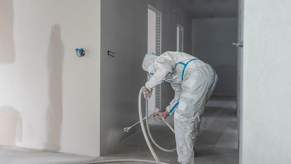 Un trabajador con un traje protector blanco rocía pintura dentro de una habitación sin terminar.