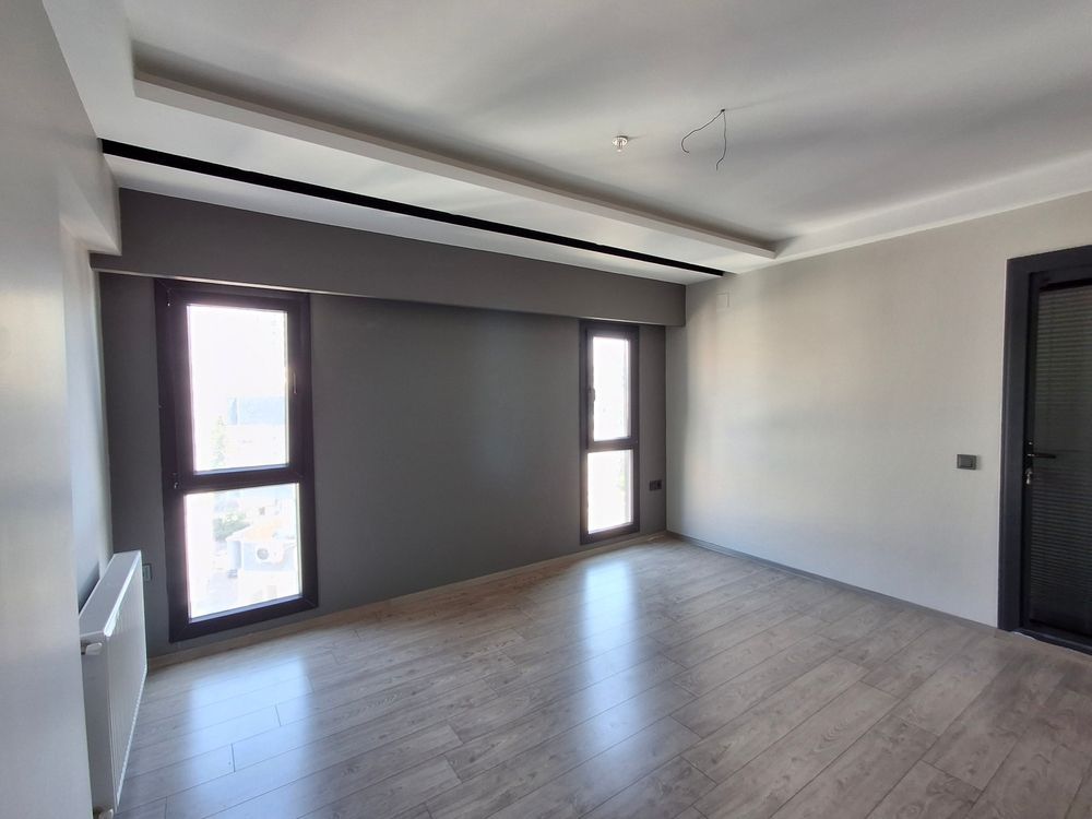 Sala de estar moderna y vacía con pared decorativa gris, grandes ventanales y suelo de madera clara.