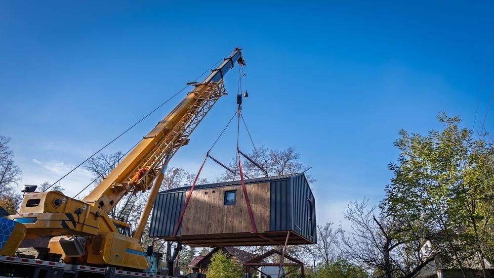 Una grúa amarilla levanta la estructura de una pequeña casa en una obra de construcción bajo un cielo azul.