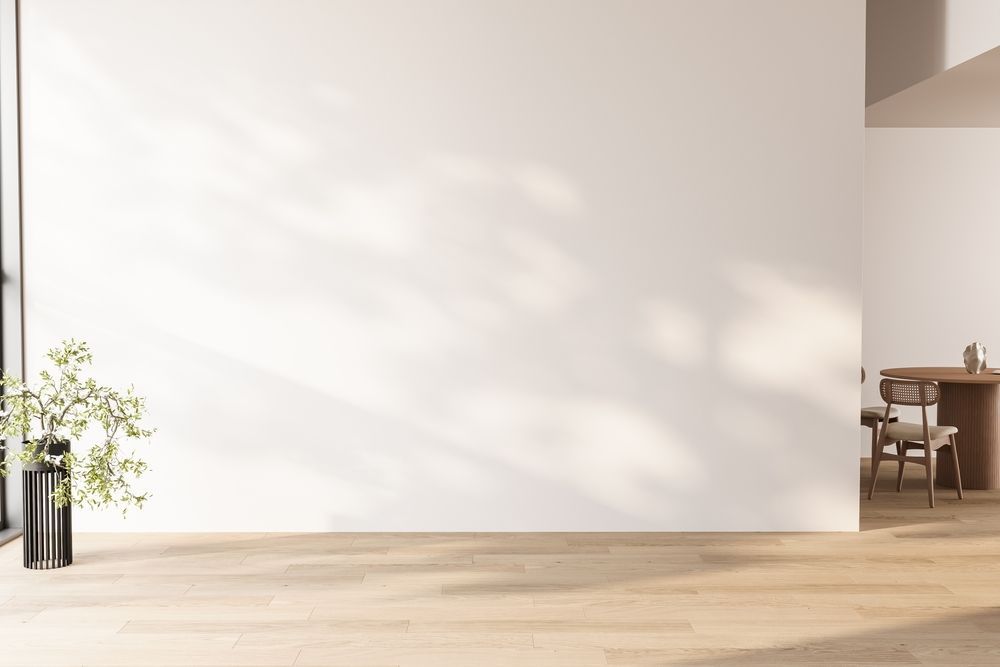 Habitación luminosa y minimalista con suelo de madera, pared blanca, una pequeña planta y un atisbo de una mesa y sillas.